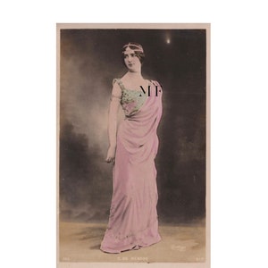 Pode incluir: Postal vintage com uma mulher em um vestido rosa esvoaçante com corpete drapeado e detalhes florais. Ela usa uma faixa de cabeça com joias e pulseiras de braço. O fundo é um gradiente escuro. O texto "C. DE MERODE" é visível.