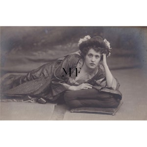 May include: Sepia-toned photograph of a person reclining on a cushion, wearing a patterned robe and a decorative hat with flowers. The person is resting their head on their hand, looking directly at the viewer.