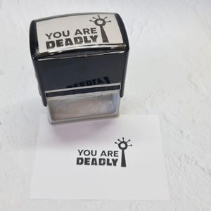 May include: A black self-inking stamp with the text "You are Deadly" and a stylized image of a hand with a single finger pointing up. The stamp is on a white surface.