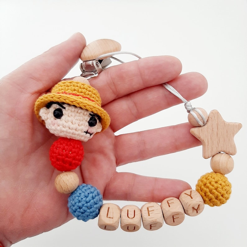 May include: A crocheted baby teether with a wooden star, wooden beads, and a crocheted character with a straw hat. The teether is spelled "LUFFY" with wooden beads.