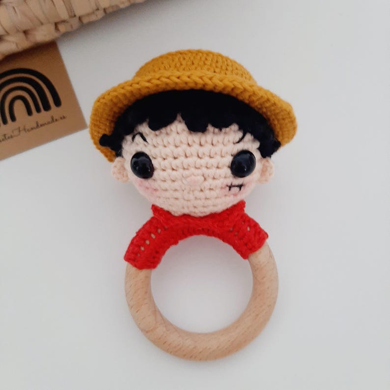 May include: A crocheted baby rattle with a wooden ring. The rattle is shaped like a person wearing a red shirt and a yellow hat. The person has black hair and brown eyes.