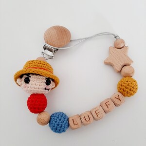 May include: A crocheted character with a yellow hat and red shirt attached to a wooden baby teether with the name "LUFFY" spelled out on it. The teether also includes a wooden star and beads.