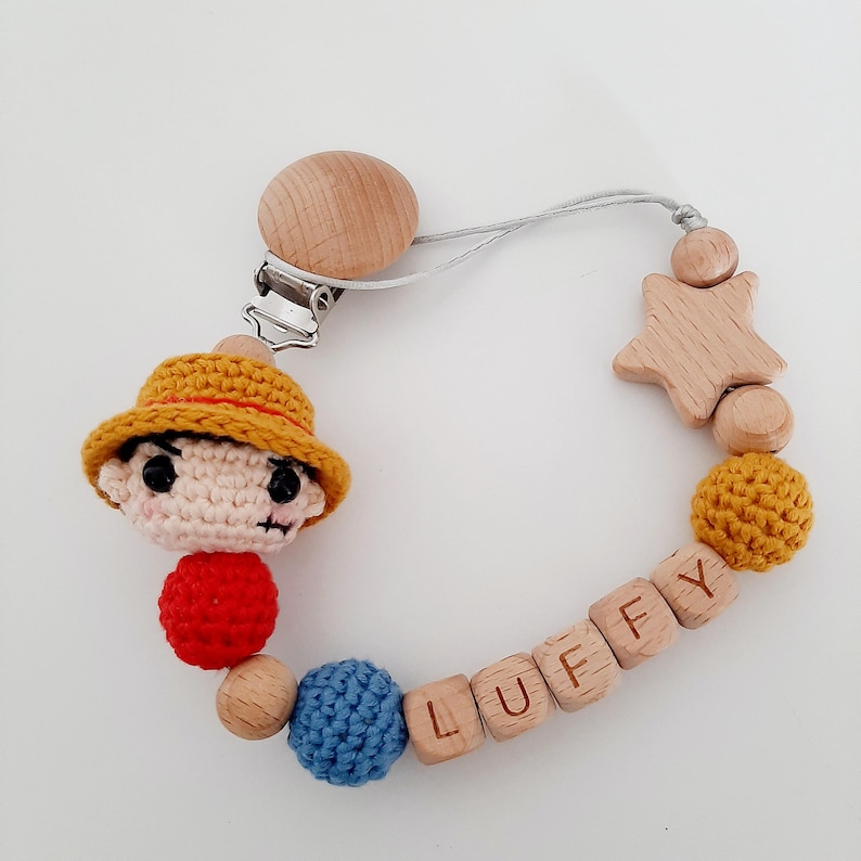 May include: A crocheted character with a yellow hat and red shirt attached to a wooden baby teether with the name "LUFFY" spelled out on it. The teether also includes a wooden star and beads.