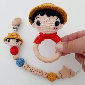 May include: A crocheted baby toy set featuring a character with a straw hat, a wooden teether ring, and a wooden star. The set also includes a pacifier clip with a crocheted character, wooden beads, and a name tag that reads "LUFFY".