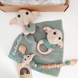 Op de afbeelding: Een groene muslin babydeken met een gehaakt bruin en groen Dobby de huiseльф speelgoed, een houten bijtring met een gehaakt groen Dobby de huiseльф speelgoed, een houten clip met een gehaakt bruin en groen Dobby de huiseльф speelgoed, en een houten kralenketting met de naam "Valentin" gespeld in houten letters.