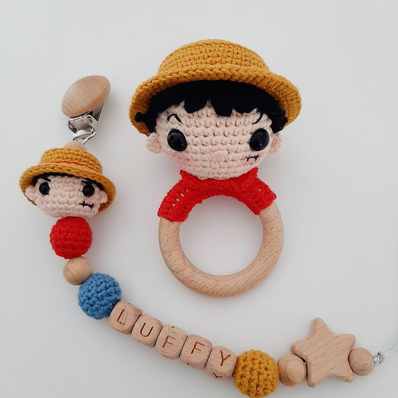 May include: A crocheted baby toy set featuring a character with a straw hat and a wooden ring rattle. The set also includes a matching character with a straw hat, a wooden star, and a beaded chain with the name "LUFFY" spelled out in wooden blocks.