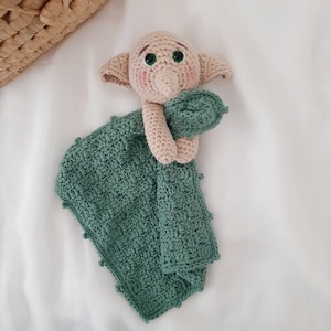 Könnte beinhalten: Eine gehäkelte grüne und beige Babydecke mit einer gehäkelten Elfenpuppe, die die Decke hält. Der Elf hat grüne Augen und eine rosa Nase.