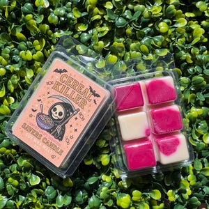 May include: Two clear plastic containers of scented soy wax melts. One has a label with the text "CEREAL KILLER" and a cartoon grim reaper. The other contains pink and white wax cubes. The background is green.