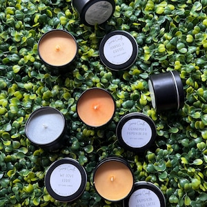May include: A collection of small, black tin candles with labels, including "Hansel & Gretel," "We Love Eddie," "Grandma's Pumpkin Pie," and "Pumpkin Spice Latte." The candles are displayed on green foliage, with visible wicks and labels.