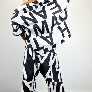 May include: A black and white patterned pantsuit with a black cap. The trousers are wide-legged and the shirt has a loose fit. The pattern is made up of large, bold letters.