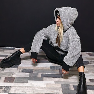 Puede incluir: Una mujer con una sudadera gris con capucha, un gorro negro y pantalones negros. Está sentada en un suelo de madera con las piernas cruzadas y lleva botas negras de tobillo.