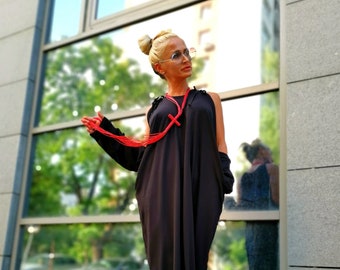 May include: A woman wearing a black one-shoulder maxi dress with a red beaded necklace and a cross pendant. She is wearing black platform sandals and has her hair pulled back in a bun.