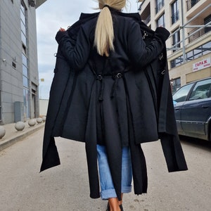 Pode incluir: Um casaco preto de manga comprida com capuz e um design longo e fluido. O casaco tem um corte assim&eacute;trico &uacute;nico e apresenta uma s&eacute;rie de al&ccedil;as e fivelas. A modelo est&aacute; usando jeans azul e sapatos pretos.