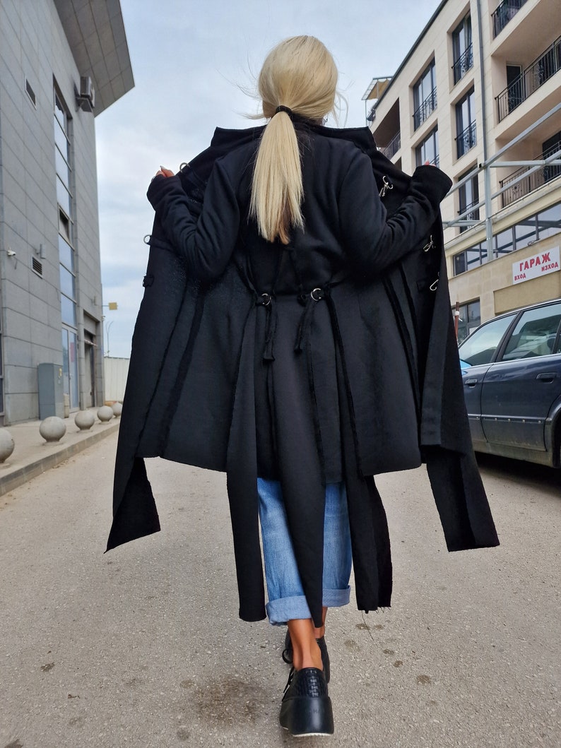 Pode incluir: Um casaco preto de manga comprida com capuz e um design longo e fluido. O casaco tem um corte assim&eacute;trico &uacute;nico e apresenta uma s&eacute;rie de al&ccedil;as e fivelas. A modelo est&aacute; usando jeans azul e sapatos pretos.