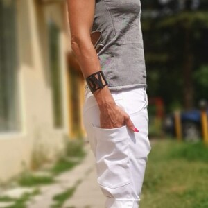 K&ouml;nnte beinhalten: Eine Frau tr&auml;gt ein graues, schulterfreies Oberteil mit einer wei&szlig;en, umgeschlagenen Shorts. Sie tr&auml;gt eine Sonnenbrille und ein schwarzes Armband mit dem Wort "LOVE" darauf. Sie steht auf einem B&uuml;rgersteig mit gr&uuml;nem Gras im Hintergrund.