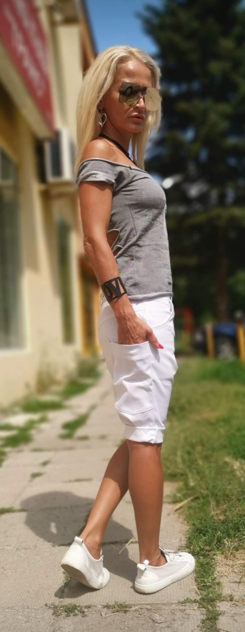 K&ouml;nnte beinhalten: Eine Frau tr&auml;gt ein graues, schulterfreies Oberteil mit einer wei&szlig;en, umgeschlagenen Shorts. Sie tr&auml;gt eine Sonnenbrille und ein schwarzes Armband mit dem Wort "LOVE" darauf. Sie steht auf einem B&uuml;rgersteig mit gr&uuml;nem Gras im Hintergrund.