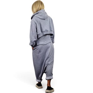 May include: A heather gray jumpsuit featuring a cropped hoodie top with long sleeves and a drop-crotch bottom. The garment has a horizontal seam across the back waist, side pockets, and cuffed hems at the ankles, paired with beige slip-on shoes.
