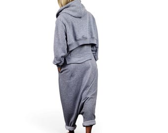 May include: A heather gray jumpsuit featuring a cropped hoodie top with long sleeves and a drop-crotch bottom. The garment has a horizontal seam across the back waist, side pockets, and cuffed hems at the ankles, paired with beige slip-on shoes.