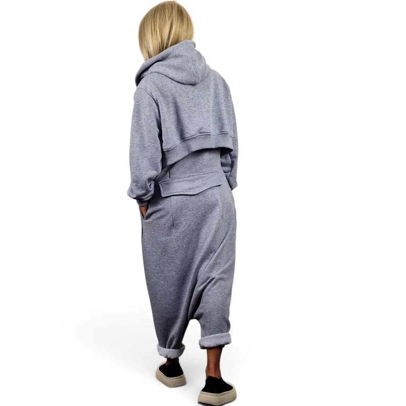 May include: A heather gray jumpsuit featuring a cropped hoodie top with long sleeves and a drop-crotch bottom. The garment has a horizontal seam across the back waist, side pockets, and cuffed hems at the ankles, paired with beige slip-on shoes.