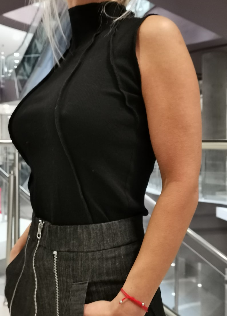 May include: A black mock neck sleeveless shirt with a seam down the front. The shirt is paired with dark gray denim pants.