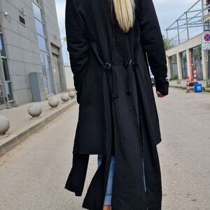 Pode incluir: Um casaco preto de manga comprida com gola alta e um design longo e fluido. O casaco tem um detalhe de n&oacute; &uacute;nico nas costas e &eacute; usado com jeans azul e sapatos de plataforma pretos.