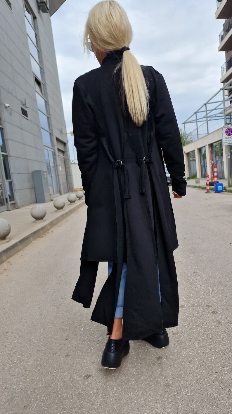 Pode incluir: Um casaco preto de manga comprida com gola alta e um design longo e fluido. O casaco tem um detalhe de n&oacute; &uacute;nico nas costas e &eacute; usado com jeans azul e sapatos de plataforma pretos.