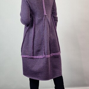 Pode incluir: Um casaco comprido roxo com um design &uacute;nico. O casaco apresenta um tecido texturizado com acabamentos roxos contrastantes ao longo das costuras e da bainha. A modelo usa cal&ccedil;as pretas e sapatos pretos.