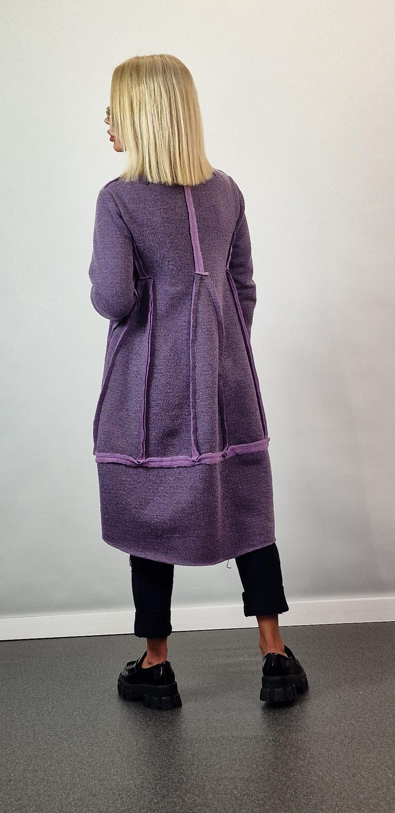 Pode incluir: Um casaco comprido roxo com um design &uacute;nico. O casaco apresenta um tecido texturizado com acabamentos roxos contrastantes ao longo das costuras e da bainha. A modelo usa cal&ccedil;as pretas e sapatos pretos.