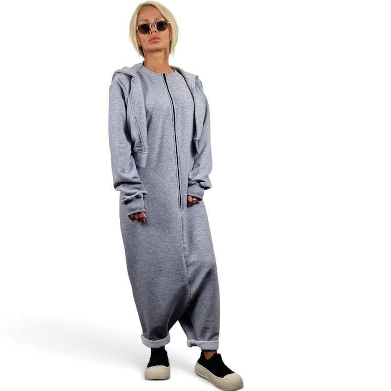 May include: A light gray long-sleeved jumpsuit featuring a full-length front zipper closure, a round neckline, and cuffed ankles. The garment is paired with a matching light gray hooded zip-up jacket worn over the top, and black sneakers.
