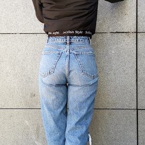 May include: A woman wearing a black puffer jacket with a hood, blue jeans, and black ankle boots. The jacket has a white elastic waistband with the text "selfish Style" printed on it.