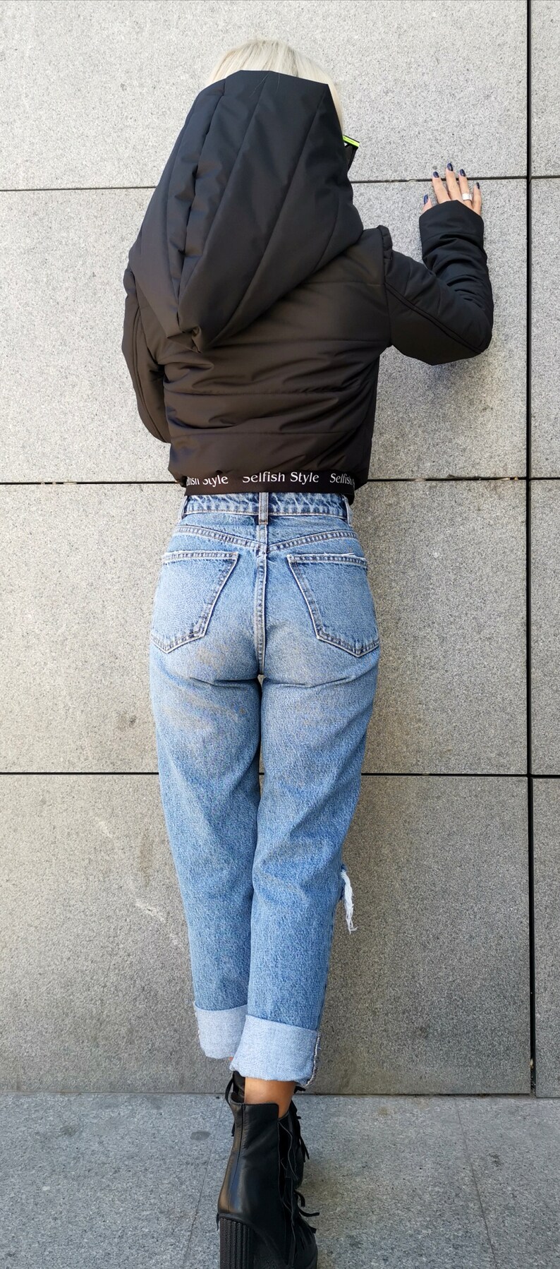 May include: A woman wearing a black puffer jacket with a hood, blue jeans, and black ankle boots. The jacket has a white elastic waistband with the text "selfish Style" printed on it.