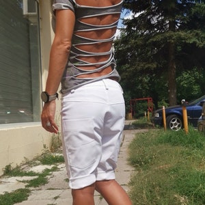 K&ouml;nnte beinhalten: Eine Frau tr&auml;gt ein graues Shirt mit Ausschnitten im R&uuml;cken und wei&szlig;e Shorts. Sie geht auf einem B&uuml;rgersteig mit gr&uuml;nem Gras auf beiden Seiten.