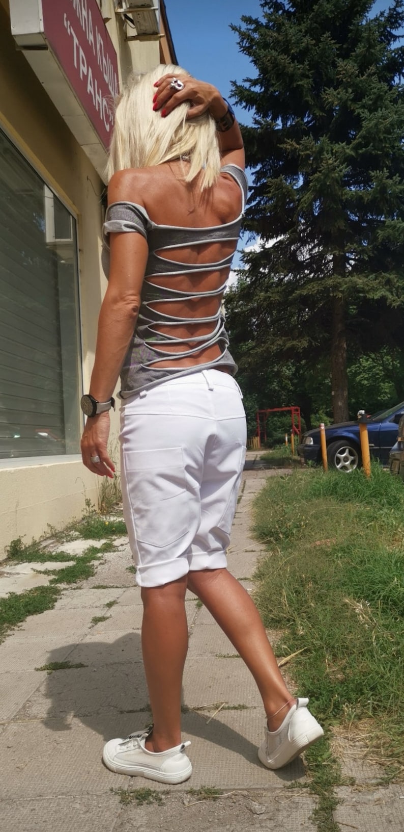 K&ouml;nnte beinhalten: Eine Frau tr&auml;gt ein graues Shirt mit Ausschnitten im R&uuml;cken und wei&szlig;e Shorts. Sie geht auf einem B&uuml;rgersteig mit gr&uuml;nem Gras auf beiden Seiten.