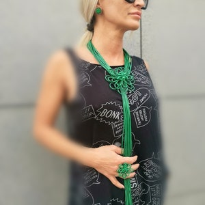 Könnte beinhalten: Eine Frau trägt ein schwarzes Kleid mit weißem Text und eine grüne Halskette mit Blumenmuster. Sie trägt auch grüne Ohrringe und einen grünen Ring.