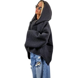 Peut inclure: Une veste à capuche zippée gris anthracite foncé avec un motif quadrillé texturé, des manches larges et un ourlet asymétrique, associée à un jean en denim bleu clair délavé présentant des déchirures et des bords effilochés sur les jambes.