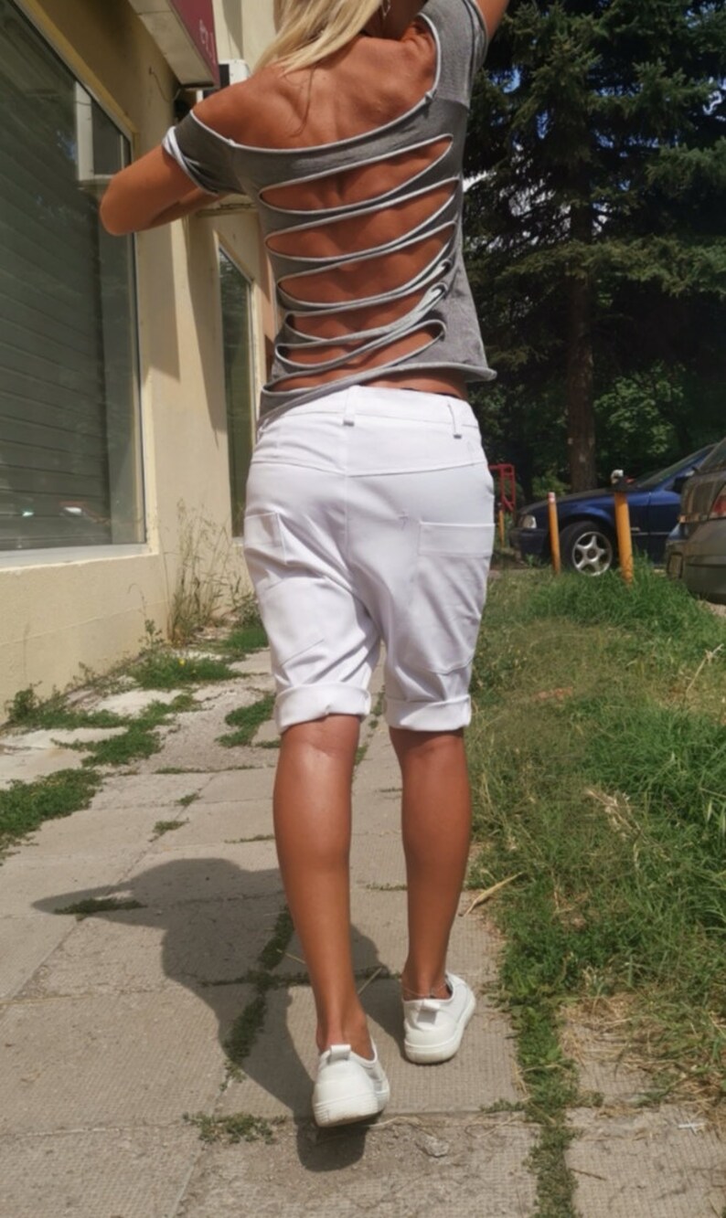 K&ouml;nnte beinhalten: Eine Frau tr&auml;gt ein graues T-Shirt mit Ausschnitten im R&uuml;cken und wei&szlig;e Shorts. Sie geht auf einem B&uuml;rgersteig.