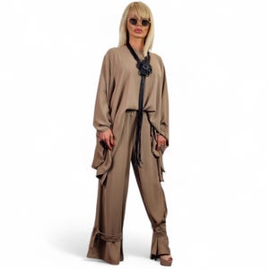 May include: A tan-colored two-piece outfit consisting of a long-sleeved top and wide-leg pants. The top has a draped design, and the pants feature a belt detail at the ankles. A black leather necklace with a floral pendant and sunglasses complete the look.