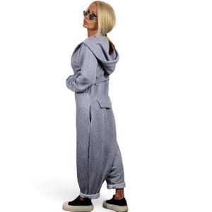 May include: A light gray hooded jumpsuit featuring long sleeves, a waist belt, and a back pocket. The garment has a relaxed fit with cuffed ankles and is paired with black sneakers. The design includes a hood and a structured waistline detail.