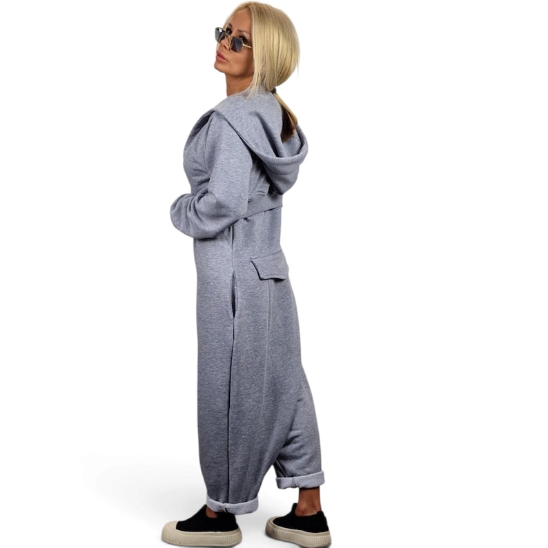 May include: A light gray hooded jumpsuit featuring long sleeves, a waist belt, and a back pocket. The garment has a relaxed fit with cuffed ankles and is paired with black sneakers. The design includes a hood and a structured waistline detail.