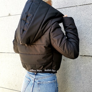 May include: Black cropped puffer jacket with a large hood and a white elastic waistband with the text "Selfish Style" repeated. The jacket is worn over blue jeans.