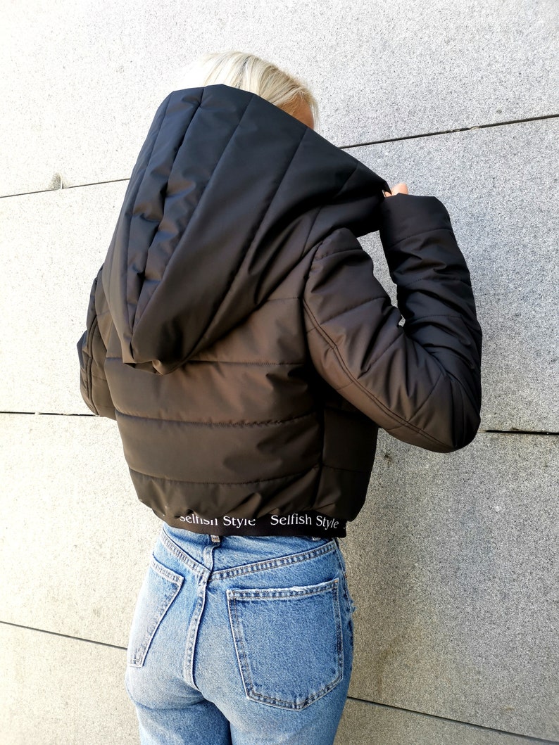 May include: Black cropped puffer jacket with a large hood and a white elastic waistband with the text "Selfish Style" repeated. The jacket is worn over blue jeans.