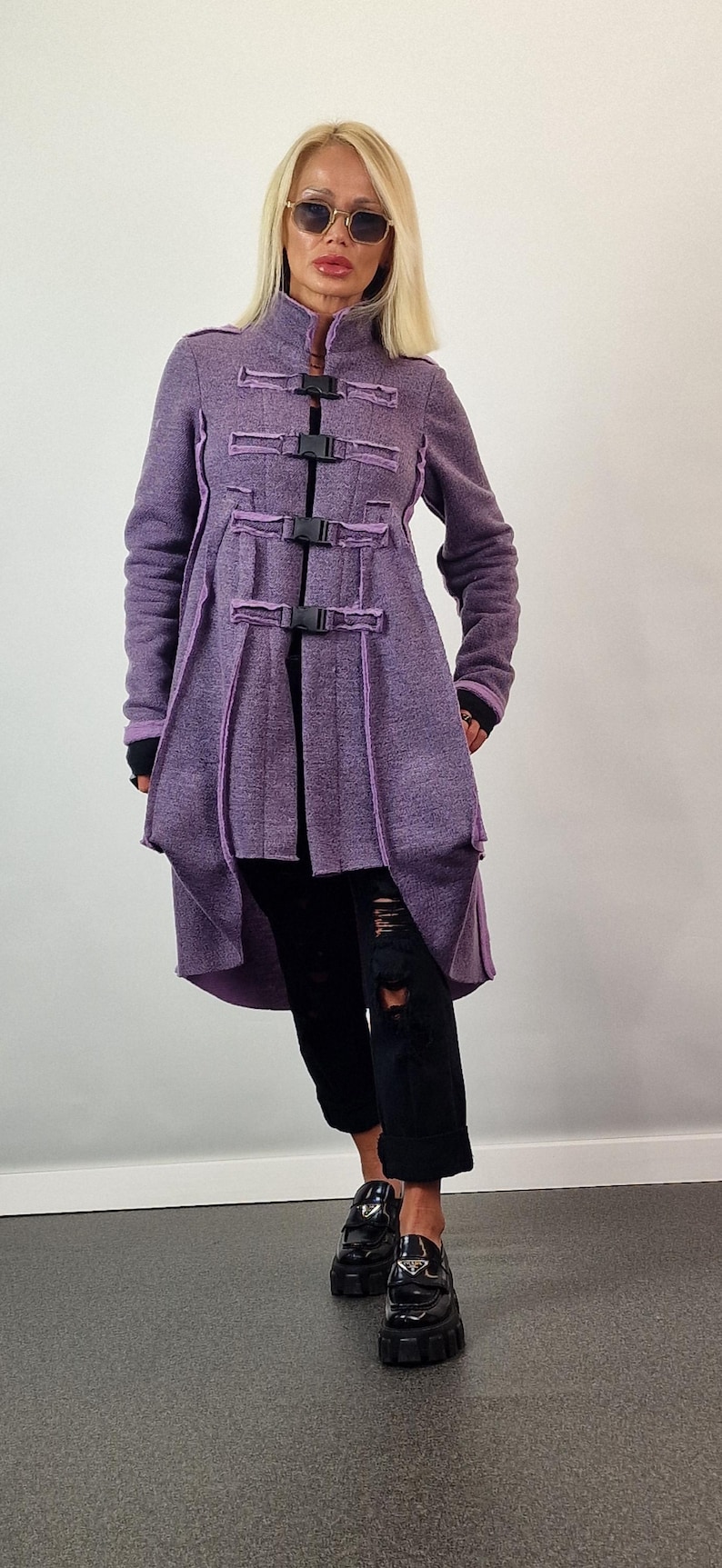 Pode incluir: Um casaco comprido roxo com fivelas e acabamentos pretos. O casaco apresenta uma gola alta e uma bainha assim&eacute;trica. A pessoa est&aacute; usando cal&ccedil;as e sapatos pretos.