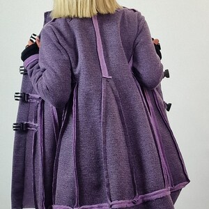 Pode incluir: Um casaco comprido, roxo, com um design &uacute;nico. O casaco apresenta uma s&eacute;rie de pain&eacute;is verticais com acabamentos contrastantes e m&uacute;ltiplos fechos de fivela. A vista traseira mostra a forma evas&eacute; do casaco e as costuras detalhadas.