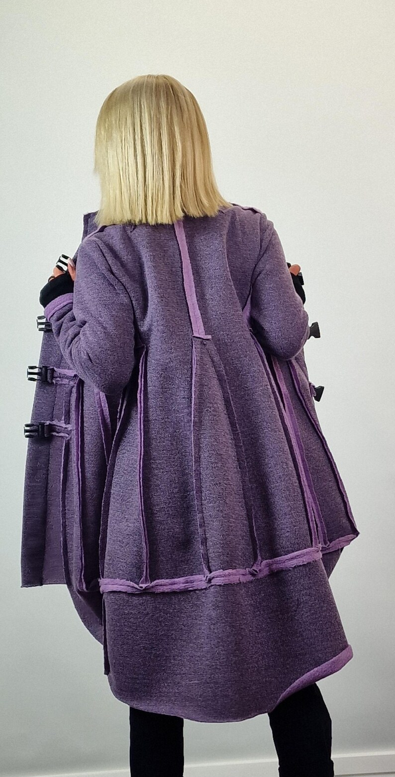 Pode incluir: Um casaco comprido, roxo, com um design &uacute;nico. O casaco apresenta uma s&eacute;rie de pain&eacute;is verticais com acabamentos contrastantes e m&uacute;ltiplos fechos de fivela. A vista traseira mostra a forma evas&eacute; do casaco e as costuras detalhadas.