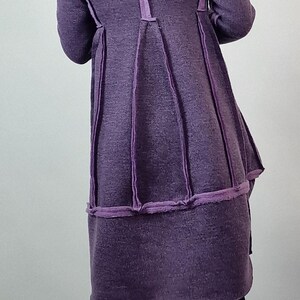 Pode incluir: Um casaco comprido roxo com um design &uacute;nico. O casaco apresenta costuras verticais e uma saia rodada. A modelo usa cal&ccedil;as pretas e sapatos pretos grossos. O casaco &eacute; feito de um tecido texturizado.