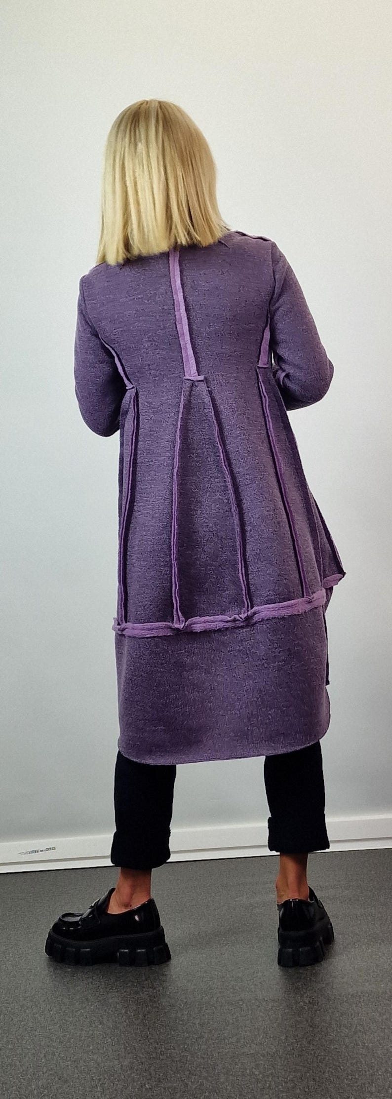 Pode incluir: Um casaco comprido roxo com um design &uacute;nico. O casaco apresenta costuras verticais e uma saia rodada. A modelo usa cal&ccedil;as pretas e sapatos pretos grossos. O casaco &eacute; feito de um tecido texturizado.