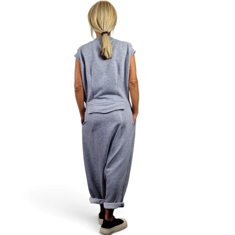May include: A sleeveless light gray jumpsuit featuring a round neckline, a horizontal flap detail across the back waist, and loose-fitting trousers with cuffed hems. The garment is made of a soft, jersey-like fabric and is shown from the back view.