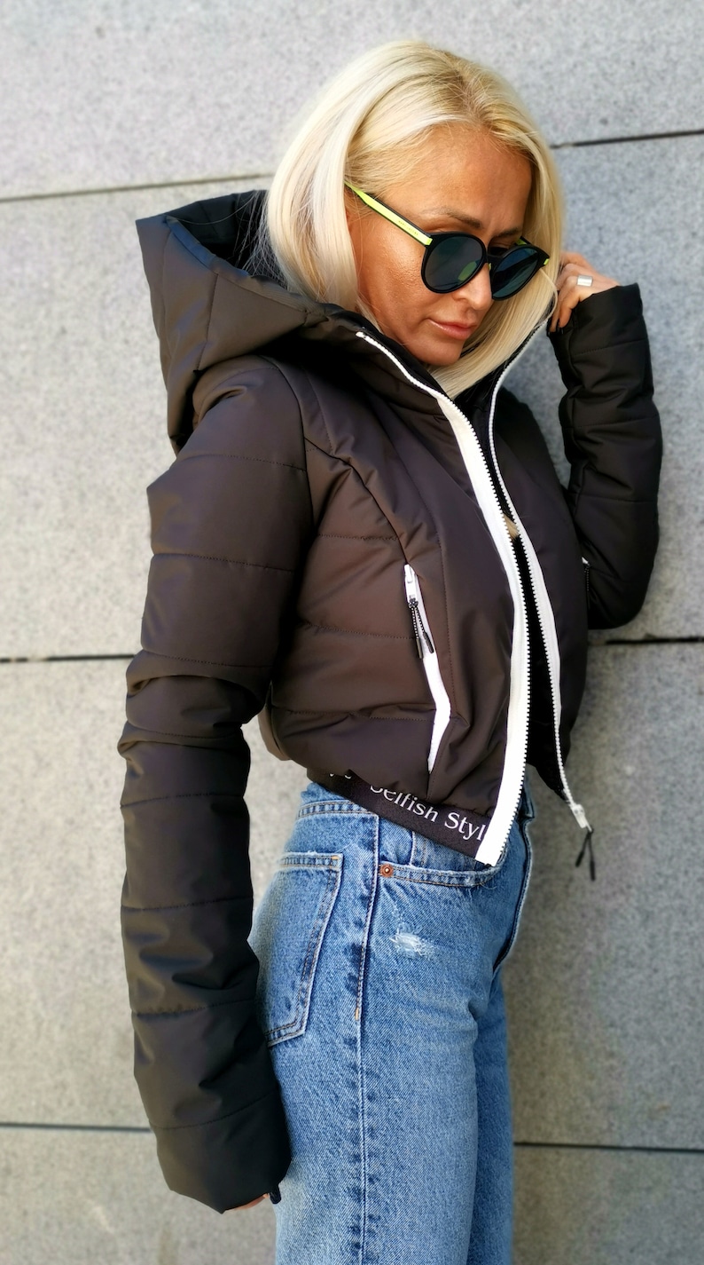 May include: A black cropped puffer jacket with white trim and a zip closure. The jacket has a hood and a small pocket on the left side. The jacket has the text "Selfish Style" printed on the bottom.