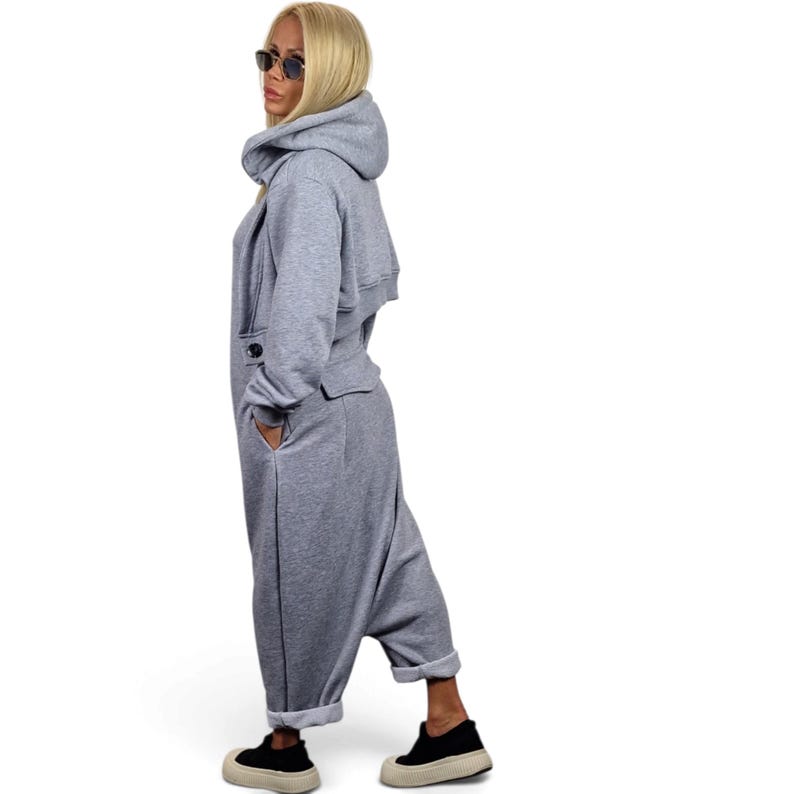 May include: A light gray hooded jumpsuit featuring a cropped overlay bodice with a single button closure at the waist, long sleeves, and loose-fitting trousers with cuffed hems. The garment is made of a soft, jersey-like fabric and includes side pockets.