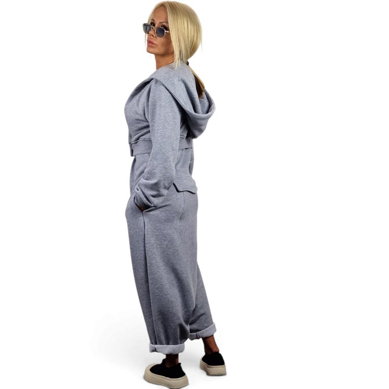 May include: A light gray hooded jumpsuit featuring long sleeves, a belted waist, and wide-leg trousers with cuffed hems. The garment is constructed from a soft, jersey-like fabric and includes a hood, providing a relaxed and comfortable fit.
